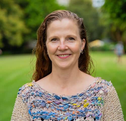 Smiling woman outdoors in park, vibrant greenery, natural light, casual summer attire, cheerful, confident, lifestyle photo.