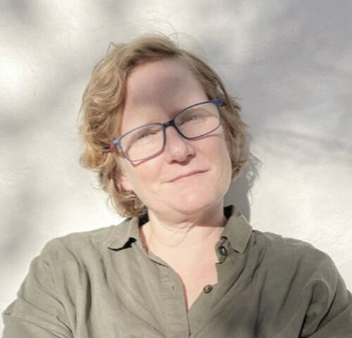 Close-up of a smiling woman with short curly hair and glasses outdoors.