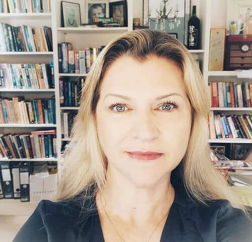 Bright woman with blonde hair taking a selfie in front of a bookshelf filled with books and decor.