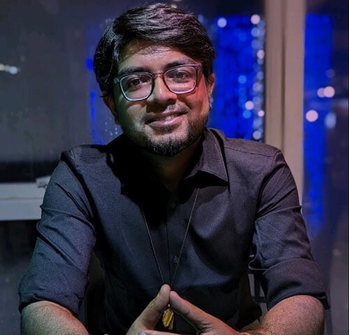 Man with glasses smiling at night indoor setting, modern city lights background.