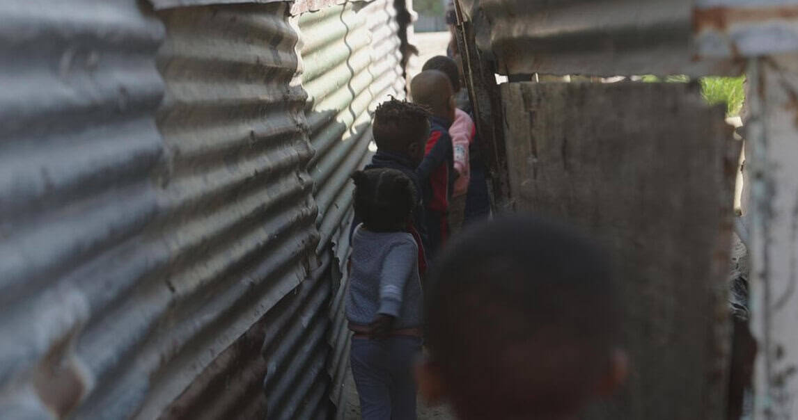 Children standing in a line between corrugated metal walls, highlighting issues of poverty and childhood hardship.